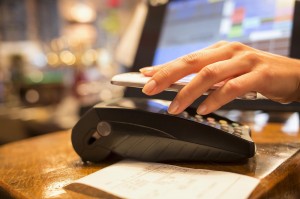A hand holds a smartphone above a card reader, making a contactless payment. A printed receipt lies on the wooden counter, and a touchscreen register is blurred in the background. The scene is in a retail or restaurant setting.