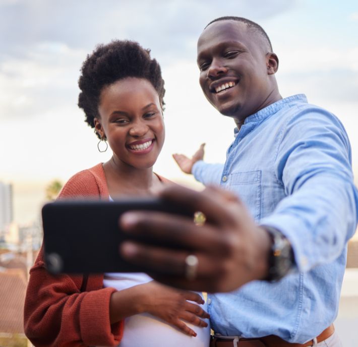 A smiling couple takes a selfie outdoors in Africa. The man, wearing a blue shirt, holds the phone and gestures with his arm, while the woman, in a rust sweater and white shirt, rests her hand on her pregnant belly under the bright African sky.