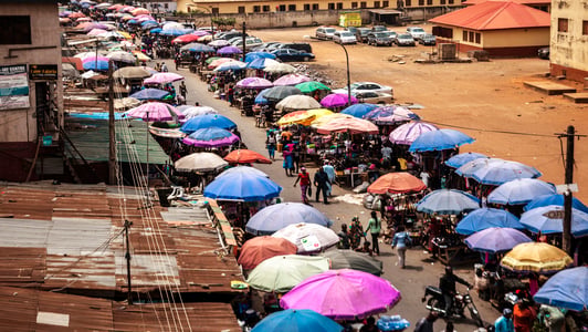 A busy outdoor market street in Africa, with colorful umbrellas covering stalls and people walking around.