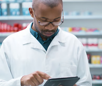 A pharmacist in a white coat uses a tablet in an African pharmacy, with shelves of medicines behind him.