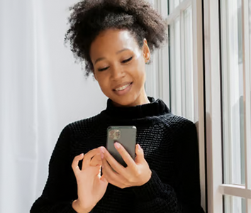 Smiling woman in a black sweater using a smartphone while standing by a window, with sunlight streaming in, capturing a moment of everyday life in Africa.