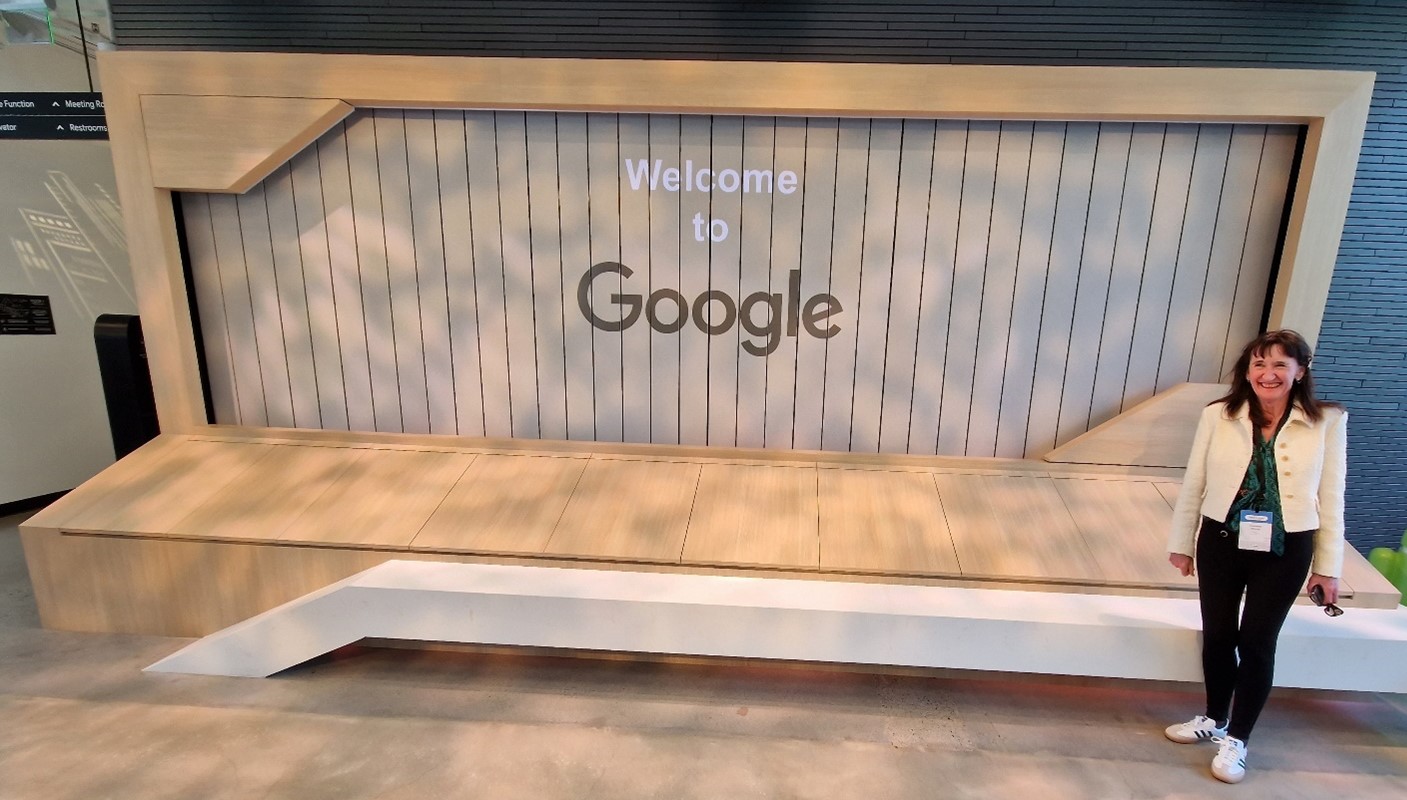 A person stands smiling in front of a large wooden welcome sign that reads "Welcome to Google." The sign is designed like a framed notice board and is set against a dark shaded wall. The person is wearing a white jacket, dark pants, and a name badge.