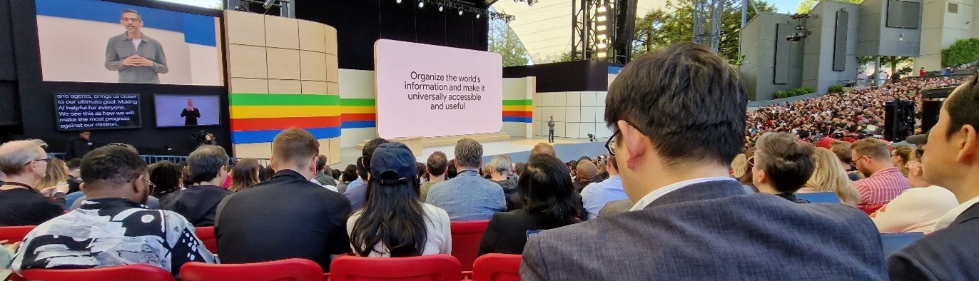 A crowded auditorium with people seated and watching a person speaking on stage. A large screen behind the speaker displays text: "Organize the world’s information and make it universally accessible and useful." Trees and buildings are visible in the background.