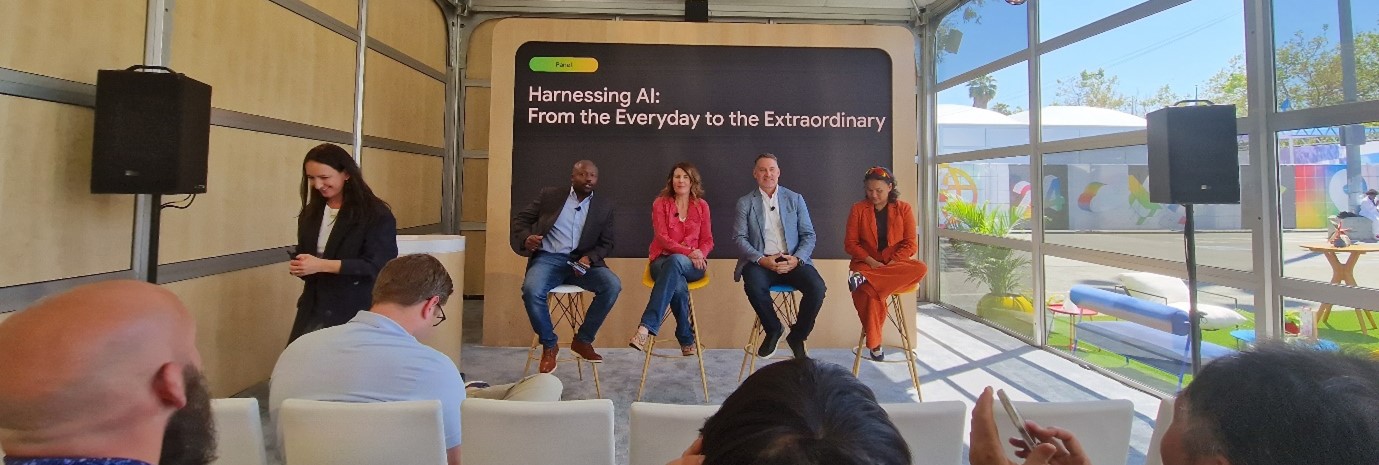 A group of five people is seated on a small stage in an outdoor tent, participating in a panel discussion titled "Harnessing AI: From the Everyday to the Extraordinary." An audience member is visible in the foreground, capturing the event on their phone.