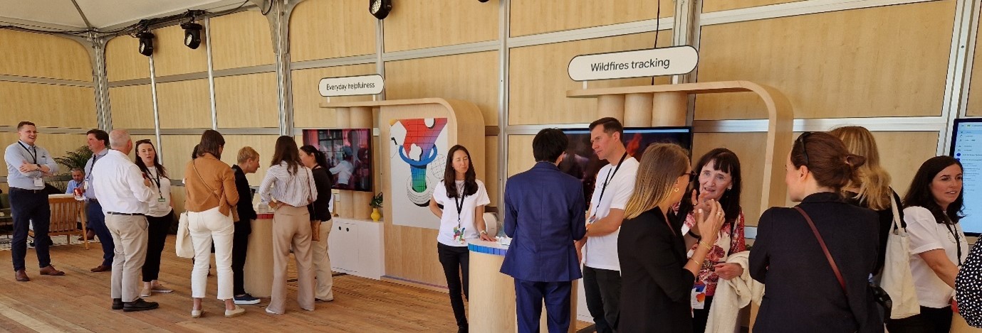 People are standing and interacting at an indoor event. There are two wooden booths labeled "Forest reporting" and "Wildfires tracking". Some individuals are conversing while others are looking at displays behind the booths.