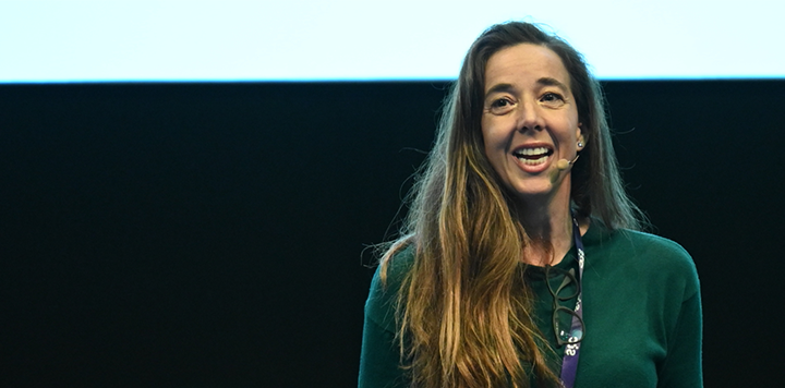 A woman with long brown hair stands speaking into a headset microphone. She is wearing a green top with a pair of glasses hanging around her neck. The background is a combination of light and dark sections.