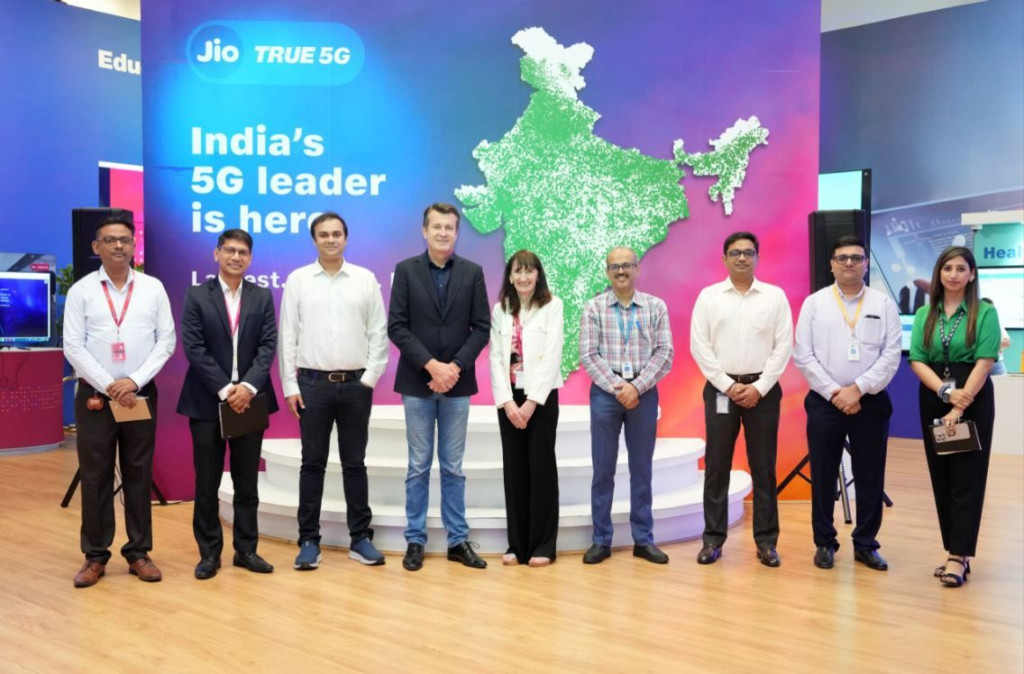 A group of nine individuals stands in front of a colorful backdrop featuring a map of India and the text "India's 5G leader is here." They are positioned in a straight line, all facing the camera. Some wear formal attire, with ID badges visible on a few.