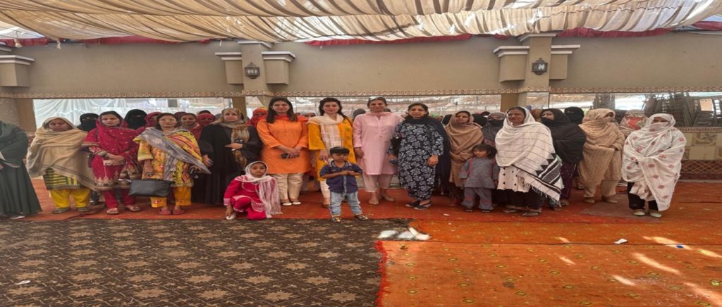 A group of around 20 people, mostly women and children, are standing in a spacious, decorated room. Some women are dressed in traditional attire. The room has draped ceilings and patterned carpets, with materials and objects scattered across the floor.