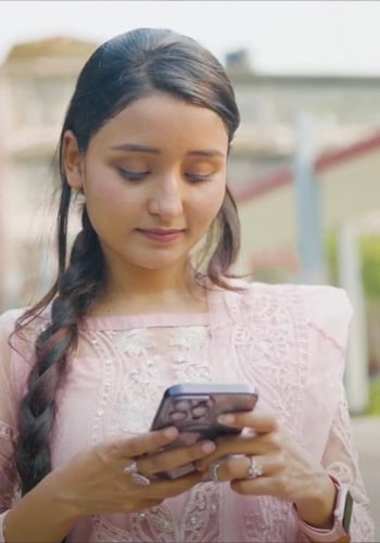 A young woman with long dark hair in a braid, wearing a light pink embroidered top, looks down and smiles slightly while using a smartphone outdoors. She is wearing a ring and a watch, with blurred buildings and greenery in the background.