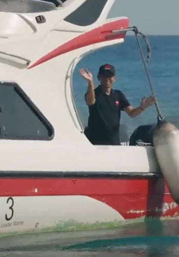 A person in a black shirt and cap stands on a white and red boat near the open cabin door, smiling and waving with one hand. The boat is on the water, with the blue sea and sky in the background. A large fender hangs beside the boat.