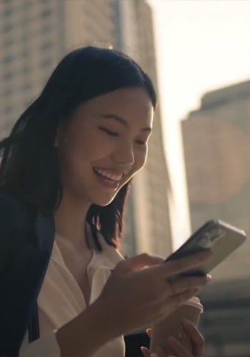 A woman with long dark hair smiles while looking at her smartphone. She is dressed in a white blouse and holding a coffee cup. Tall city buildings are blurred in the background, suggesting she is outdoors in an urban setting.