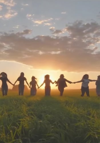 A group of eight people stand in a grassy field, holding hands in a line at sunset. The sun is low on the horizon, casting a warm glow and long shadows, with clouds scattered across the sky. The scene conveys unity and togetherness.