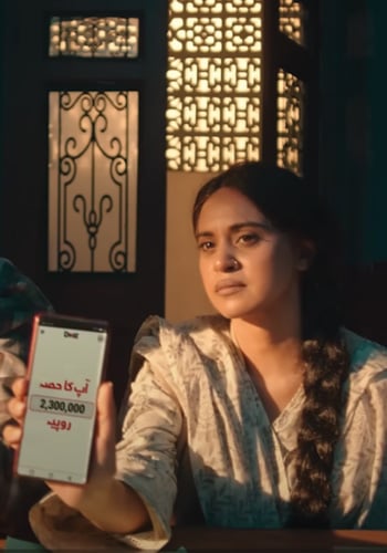 A woman with a serious expression sits indoors, holding a smartphone toward the camera. The phone screen displays Arabic text and the number “2,300,000.” Warm light from ornate windows and decorative metalwork illuminates her and the background.