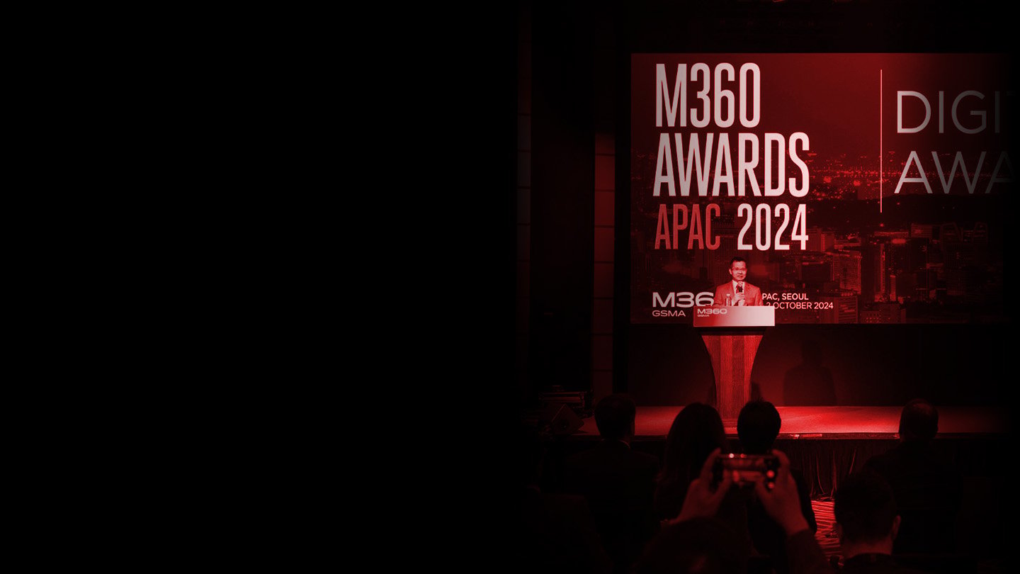 A person stands at a podium on stage, speaking at the M360 Awards APAC 2024 event. The backdrop displays the event name in large white letters on a red background. Audience members are seated, and one is taking a photo with a phone.