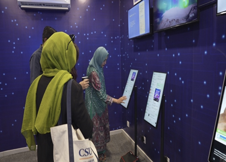 Three people stand in front of large touchscreen displays mounted on a blue wall with a dotted digital pattern. Two women in headscarves interact with the screens; one carries a white tote bag labeled "CSU." Another person stands behind them.