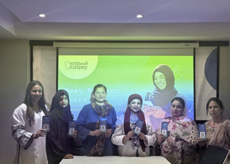 Six women stand indoors holding up pamphlets, smiling at the camera. Behind them is a projector screen displaying "Kistpay" and images of women in hijabs, with green and blue background graphics and text about digital empowerment.
