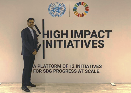 A man in a dark suit stands smiling beside a sign that reads “HIGH IMPACT INITIATIVES: A platform of 12 initiatives for SDG progress at scale.” The United Nations and Sustainable Development Goals logos are visible at the top.