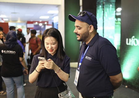 A woman and a man in navy blue polo shirts stand together at an indoor event. The woman is smiling and looking at her phone, while the man, wearing a cap and lanyard, looks on. People and display booths are visible in the background.