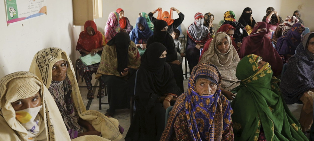 A group of women wearing colorful traditional clothing and headscarves sit closely together in a waiting room. Some look toward the camera, while others face forward or talk among themselves. The room has plain walls and a poster is visible on the left.