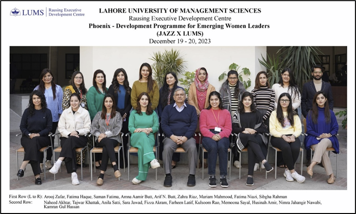 Group photo of 22 people, mostly women, seated and standing in two rows. They are posing for a formal picture at LUMS’ Phoenix women’s leadership program. A man sits in the center front. Everyone is dressed in winter attire, smiling, in a bright, indoor setting.