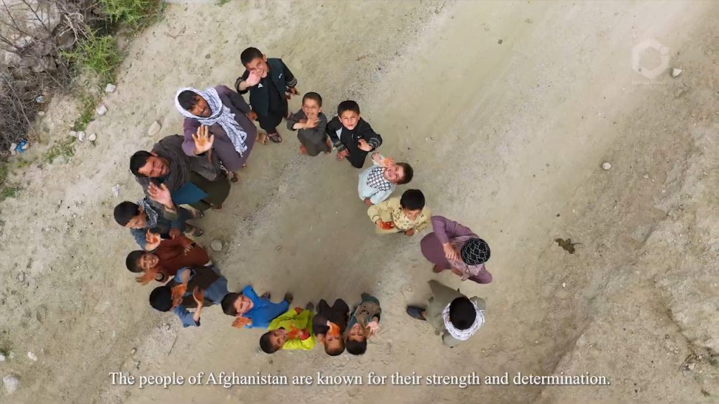 A group of Afghan children and adults stand outdoors on dusty ground, looking up and waving at the camera. Most are smiling. The photo is taken from above. The caption at the bottom reads, “The people of Afghanistan are known for their strength and determination.”.