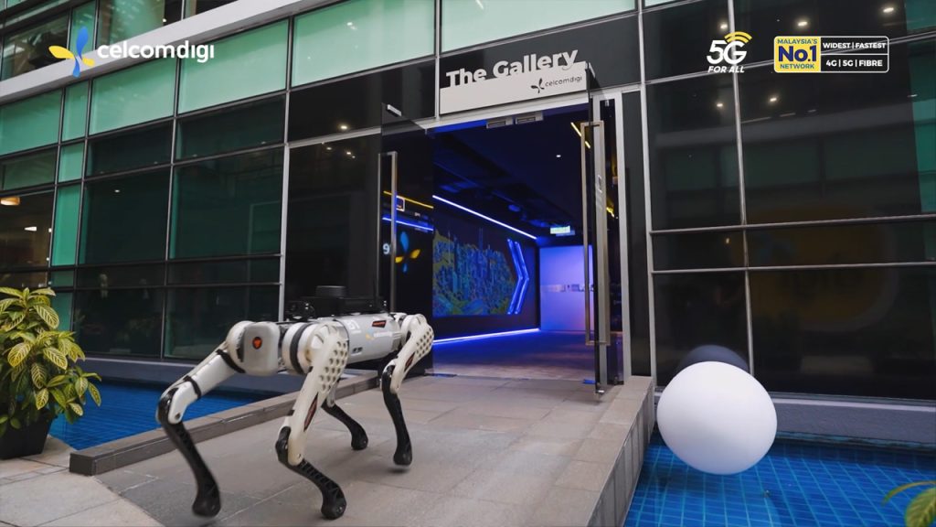 A robotic dog walks on a paved path outside a modern glass building labeled "The Gallery". Blue accent lighting glows inside, and company logos for CelcomDigi, 5G, and Malaysia's No.1 network are visible on the building facade. A round white object is near the water feature.
