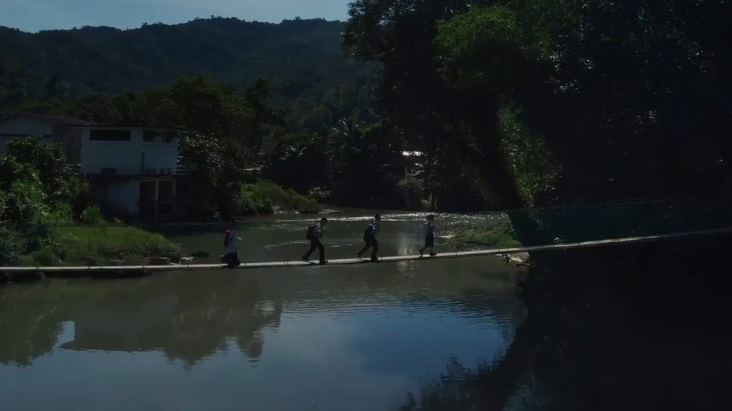 Four people walk single file along a narrow bridge over a calm river, surrounded by lush green trees and hills. A house stands near the riverbank on the left, and forested mountains rise in the background under a clear sky.