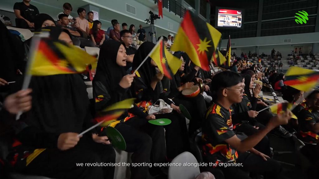 A group of fans in black and yellow sports attire wave Sarawak flags and cheer enthusiastically in a stadium. Most are wearing headscarves. A scoreboard and more spectators are visible in the background. Subtitle reads: "we revolutionised the sporting experience alongside the organiser".