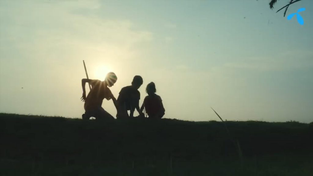 Three children are silhouetted against a sunset sky, sitting or kneeling on grassy ground. One child holds a stick, and the sun is visible behind them, creating a peaceful, nostalgic scene. A small blue logo appears in the top right corner.