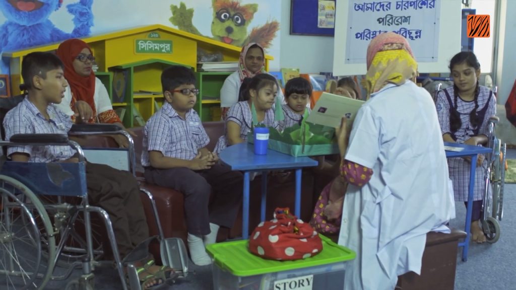 A teacher in a white headscarf reads a book to a group of young students, some in wheelchairs, in a colorful classroom. The children wear matching uniforms and listen attentively. Educational posters and toys are visible in the background.