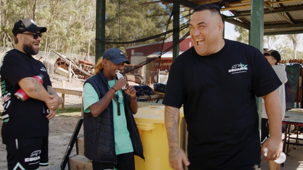 Three men stand outdoors under a metal shelter, laughing and smiling. Two wear black T-shirts and sunglasses, while the third wears a teal shirt and black vest. A yellow bin, tables, and trees are in the background, suggesting a casual, fun gathering.