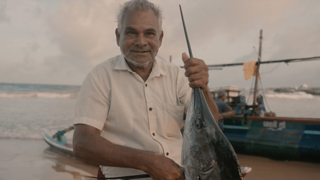 An older man with gray hair and a short beard smiles while holding a large fish, possibly a swordfish, by the head on a beach. Behind him, there are fishing boats and the sea under a cloudy sky, suggesting a coastal fishing village.