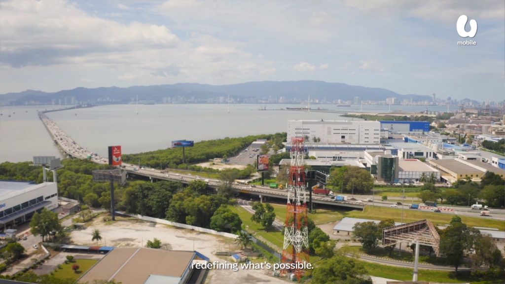 A wide aerial view of a coastal city shows a long bridge stretching over water, modern buildings, a red and white telecom tower, green trees, and distant mountains under a partly cloudy sky. Subtitles read, “redefining what’s possible.”.