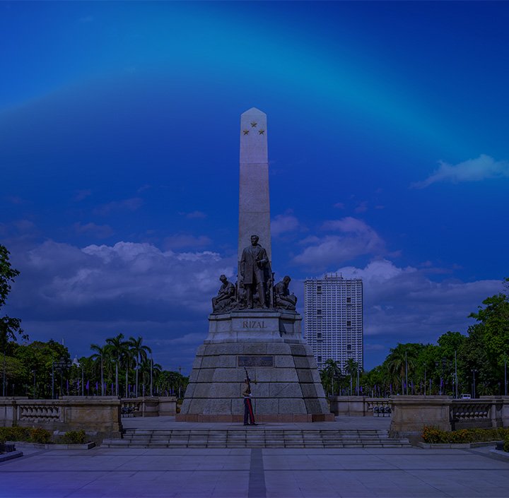 A tall stone monument with a statue of a man and several figures at its base, labelled “RIZAL,” stands in a spacious plaza. A modern high-rise and trees are in the background, under a blue sky with scattered clouds and a faint rainbow arc.