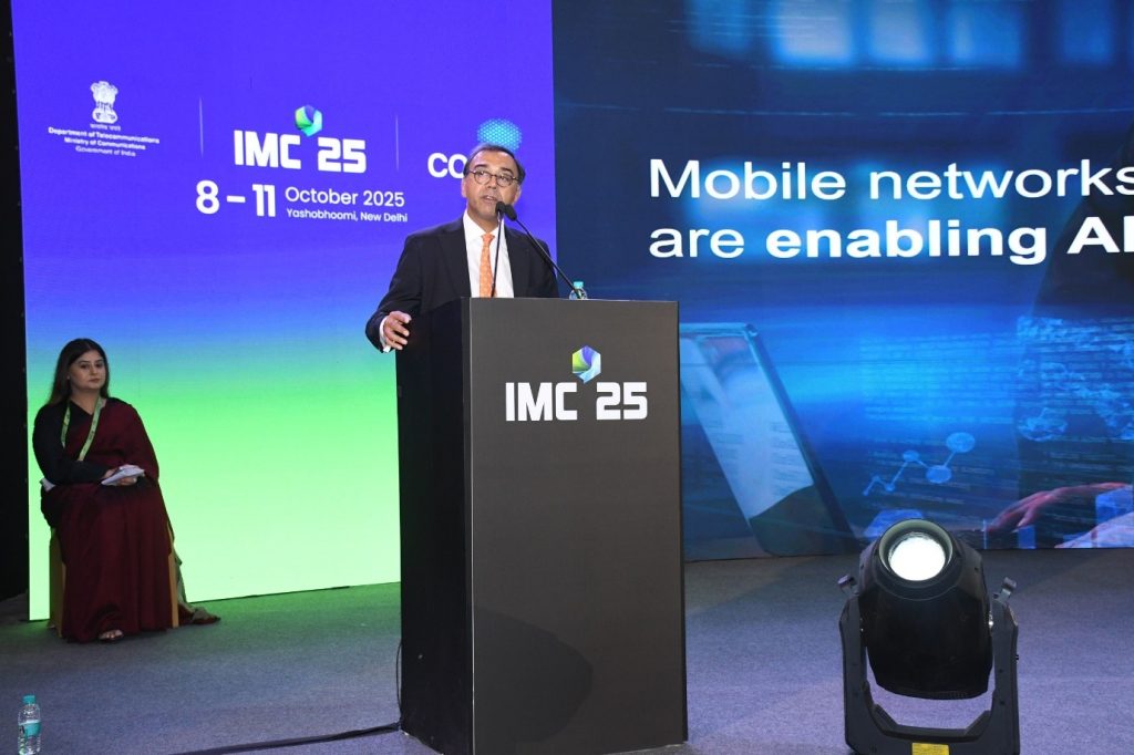 A man in a suit speaks at a podium labeled "IMC 25" on a stage. Behind him, a large screen displays event details: "IMC 25, 8-11 October 2025, Yashobhoomi, New Delhi." A seated woman in a red sari is visible to the side.