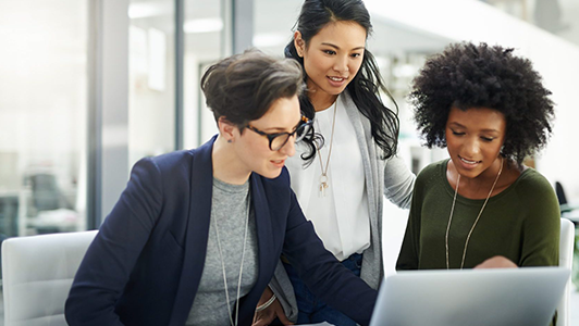 Three women collaborate around a laptop in a modern office. One woman with short hair and glasses types, while the other two, one with long hair and one with curly hair, lean in and look at the screen, all appearing engaged and focused on the task.