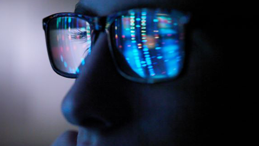 Close-up of a person wearing glasses, with blue and multicolored light patterns resembling computer code or data reflected in the lenses, suggesting focus on a digital screen or programming activity in a dimly lit environment.
