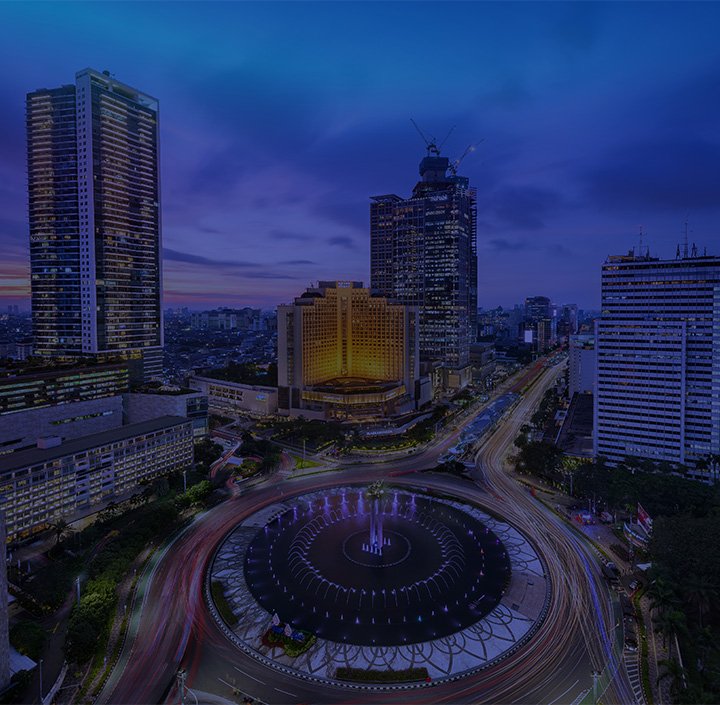 Aerial night view of Jakarta’s Bundaran HI roundabout in the Asia Pacific, with a central illuminated fountain, colorful light trails from traffic, and tall skyscrapers under a deep blue sky. The city skyline is prominent in the background.