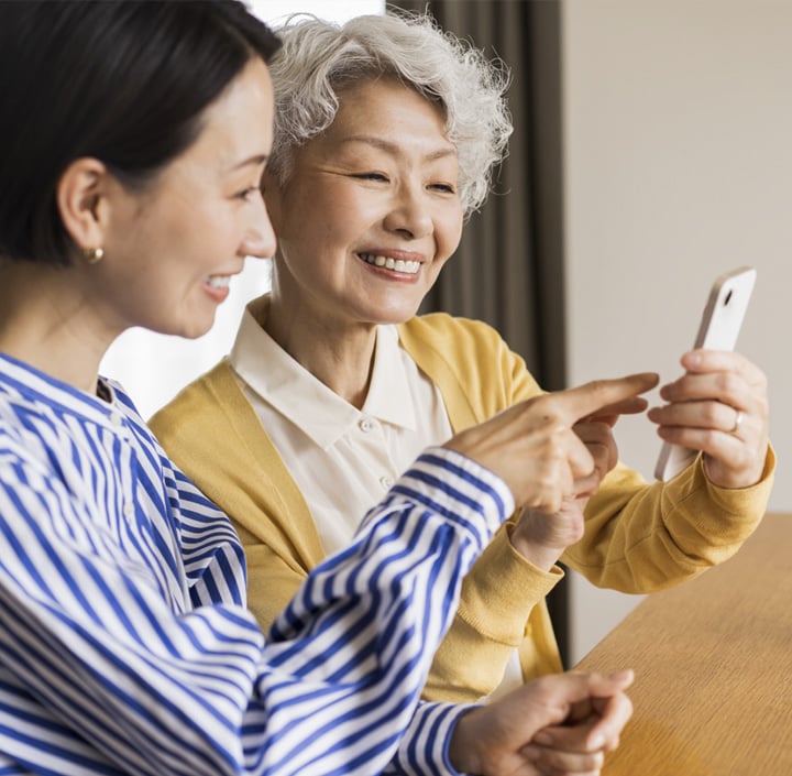 Two women in the Asia Pacific region smile and look at a smartphone together, one pointing at the screen as they share a joyful moment.
