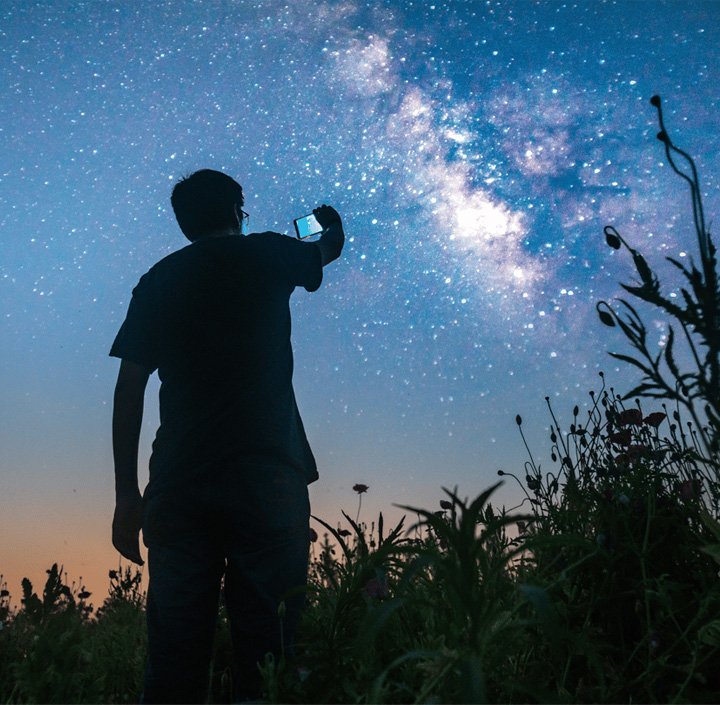 A person photographs the starry night sky and Milky Way, standing in a field with silhouetted plants in the Asia Pacific region.