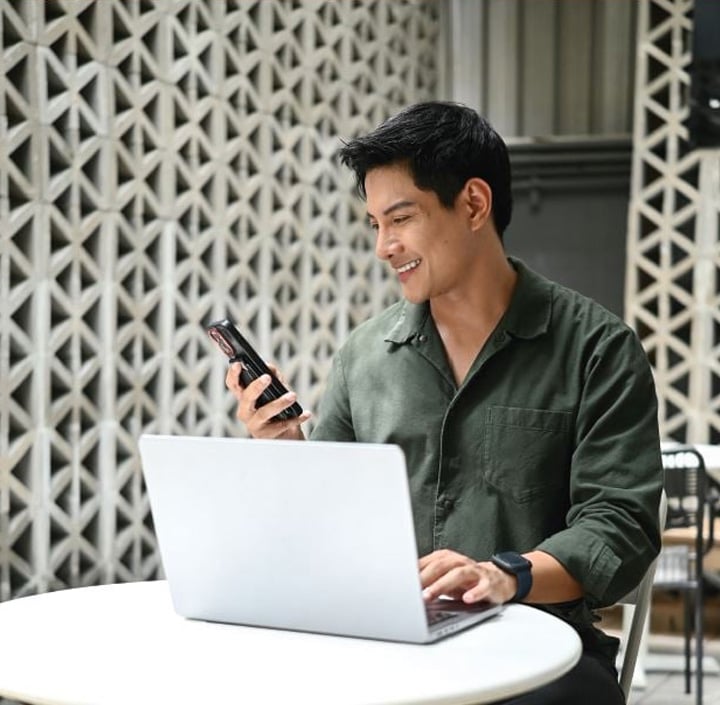 Man smiling at his phone while working on a laptop at a round white table indoors, in a modern Asia Pacific setting.