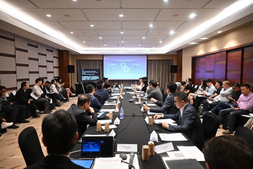 A large conference room with many people seated around a long table, listening to a presentation displayed on a screen at the front. Attendees are in business attire, some using laptops. The room has modern lighting and wood-paneled walls.