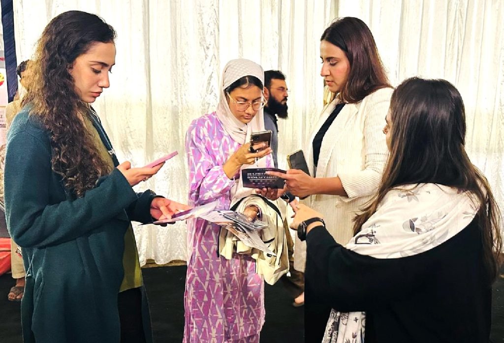 Four women stand together indoors in front of a white curtain, each focused on a phone or reading material in their hands. One woman wears a light purple patterned dress and headscarf; the others wear dark or light outerwear. A bearded man is visible in the background.