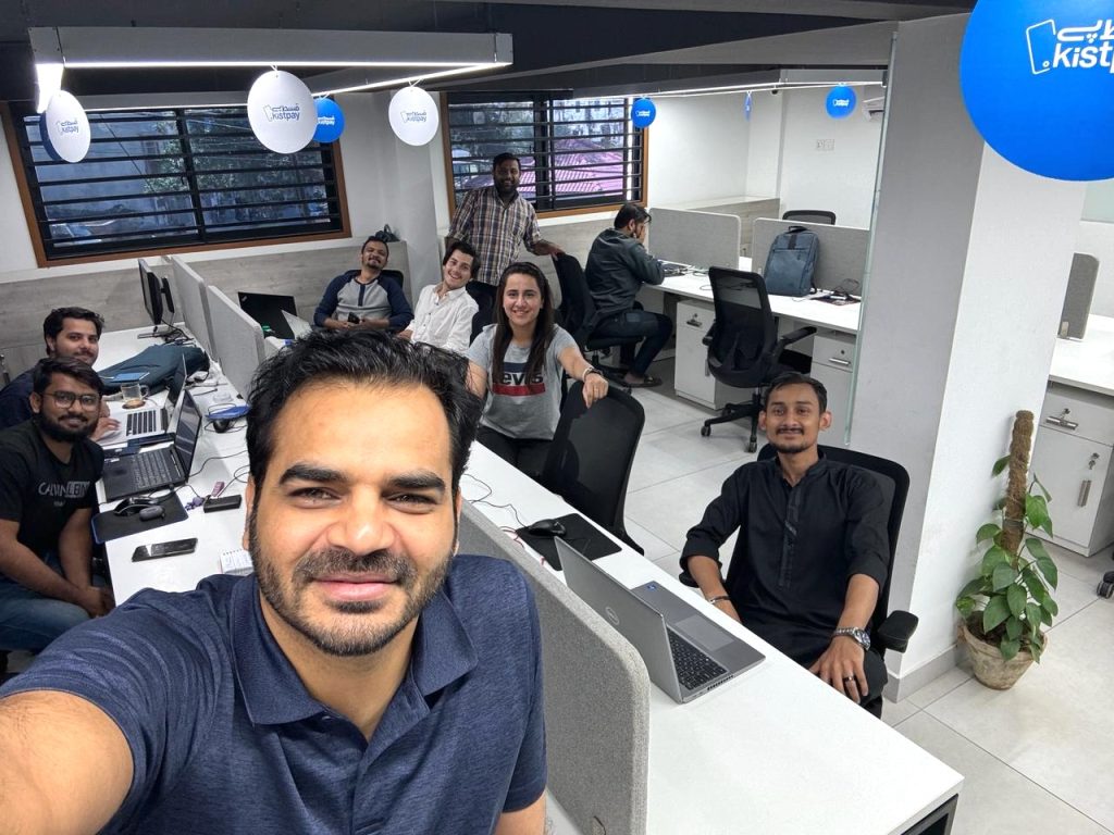 A group of eight people, some seated and some standing, smile for a selfie in a modern office with white desks, computers, blue logo signs, and large windows. One man in front holds the camera. The workspace is bright and organized.