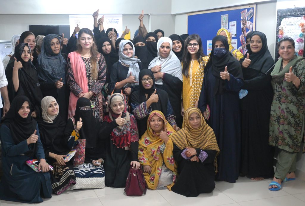 A group of women, many in black or patterned hijabs and abayas, stand and kneel indoors, smiling and giving thumbs up. Some wear glasses or colorful scarves. The background features posters and a bulletin board, creating a lively, supportive atmosphere.