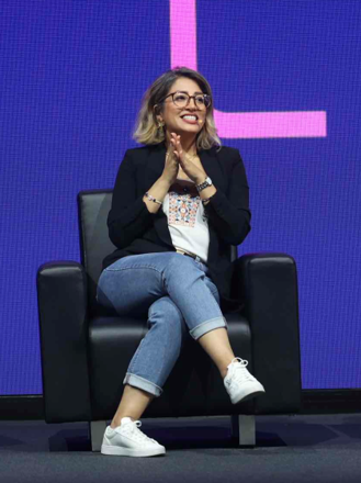 A woman with shoulder-length blonde hair, glasses, and a patterned shirt under a black blazer sits cross-legged on a black armchair, smiling with her hands clasped under her chin, against a purple background with pink geometric shapes.