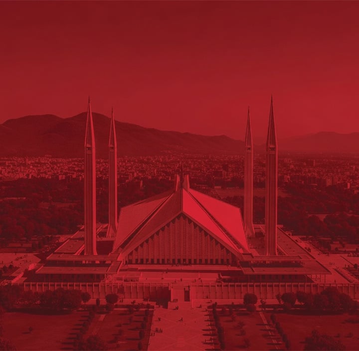 A red-tinted aerial view of the Faisal Mosque in Islamabad, Pakistan, an architectural landmark of the Asia Pacific region. The mosque features four tall minarets and a triangular roof, set against the backdrop of distant mountains and a sprawling cityscape.