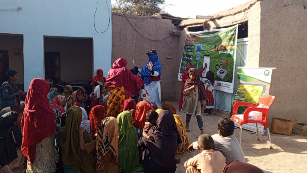 A group of women and children, many wearing colorful traditional clothing, sit and stand in a courtyard in front of mud buildings. Two women lead a discussion near posters and banners about health or sanitation. Sunlight brightens the scene.