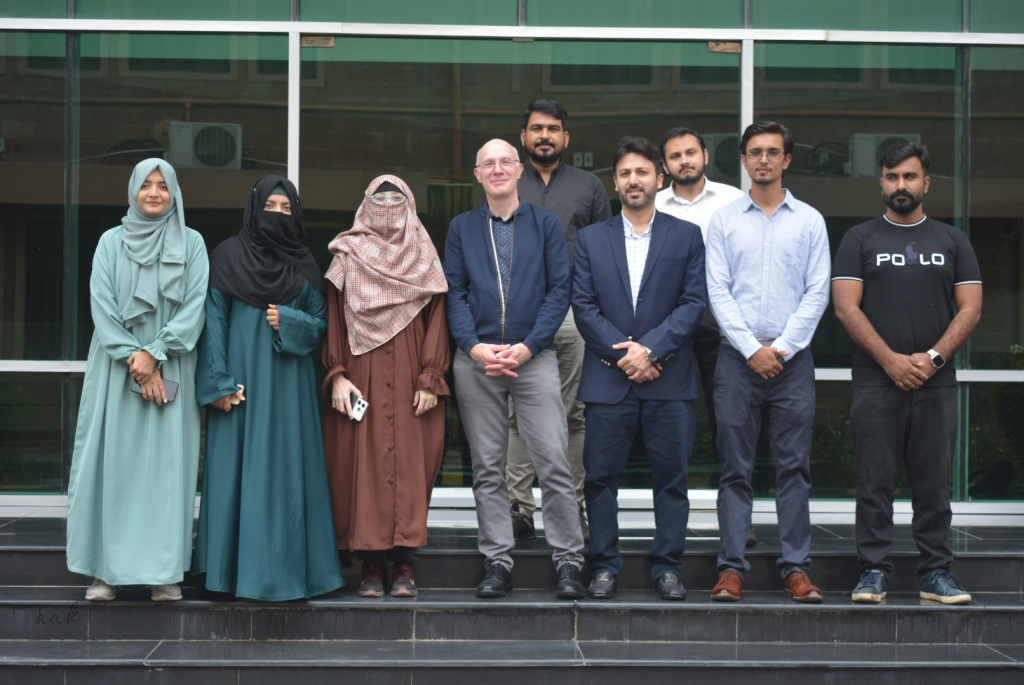 A group of eight adults, four women and four men, stand in two rows on black steps outside a building with large glass windows. The women wear modest clothing and headscarves; the men wear business or casual attire. All face the camera and smile.
