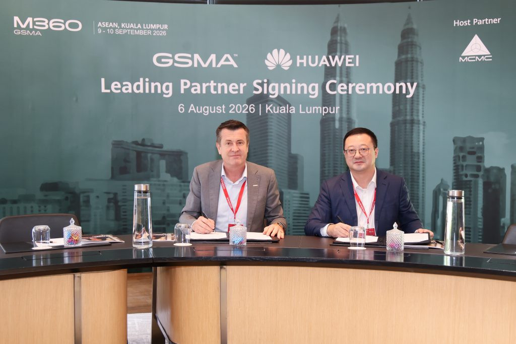 Two men in business attire sit at a table, smiling and signing documents at a GSMA and Huawei Leading Partner Signing Ceremony in Kuala Lumpur. The backdrop shows event details and images of skyscrapers. Water bottles, cups, and microphones are on the table.